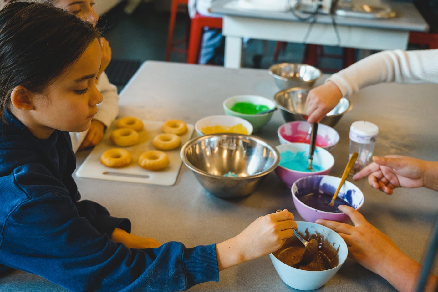 Children mixing bowls of bright icing beside a board of freshly baked doughnuts