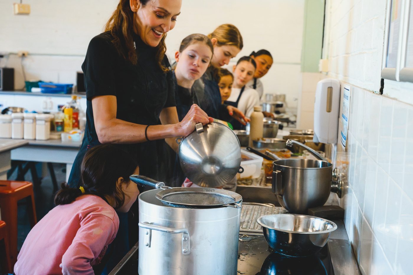 Yvonne lifting a lid at the stove with a line of students watching what is cooking
