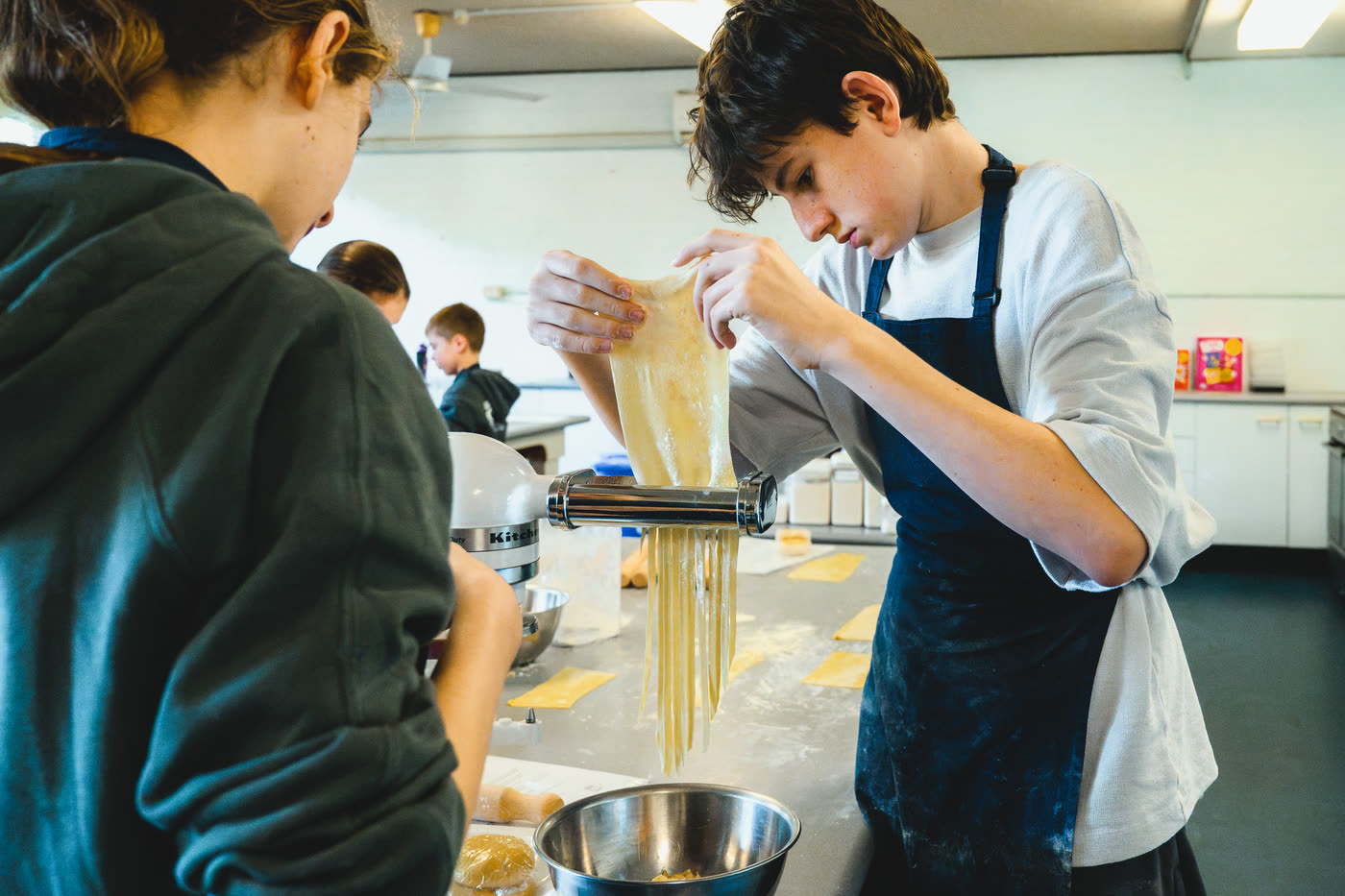 A boy feeding a sheet of pasta through the cutter while another student works at the bench beside him.