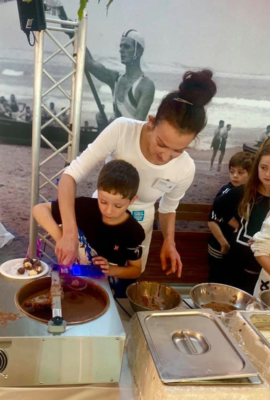 A young boy spooning melted chocolate at a tempering machine set up by the surf club, with the beach behind