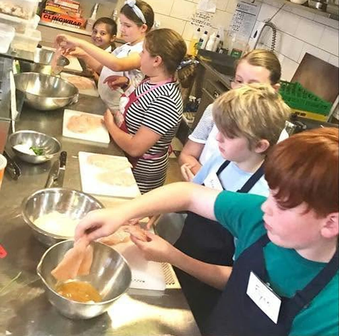 Children crowded around a bench of mixing bowls, one reaching in to fold a batter