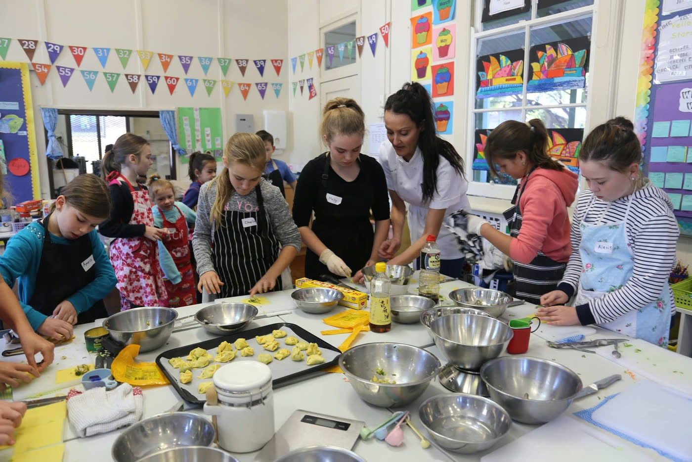 A school hall session with rows of children at tables, trays of baking in front of them