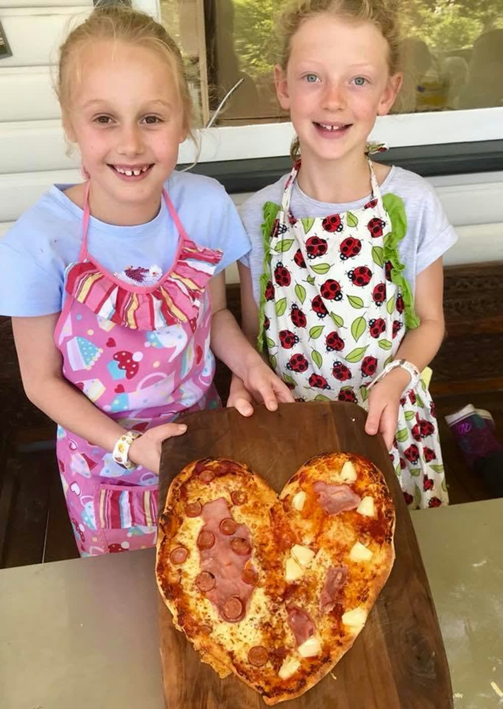 Two girls in floral aprons presenting the heart-shaped pizza they made